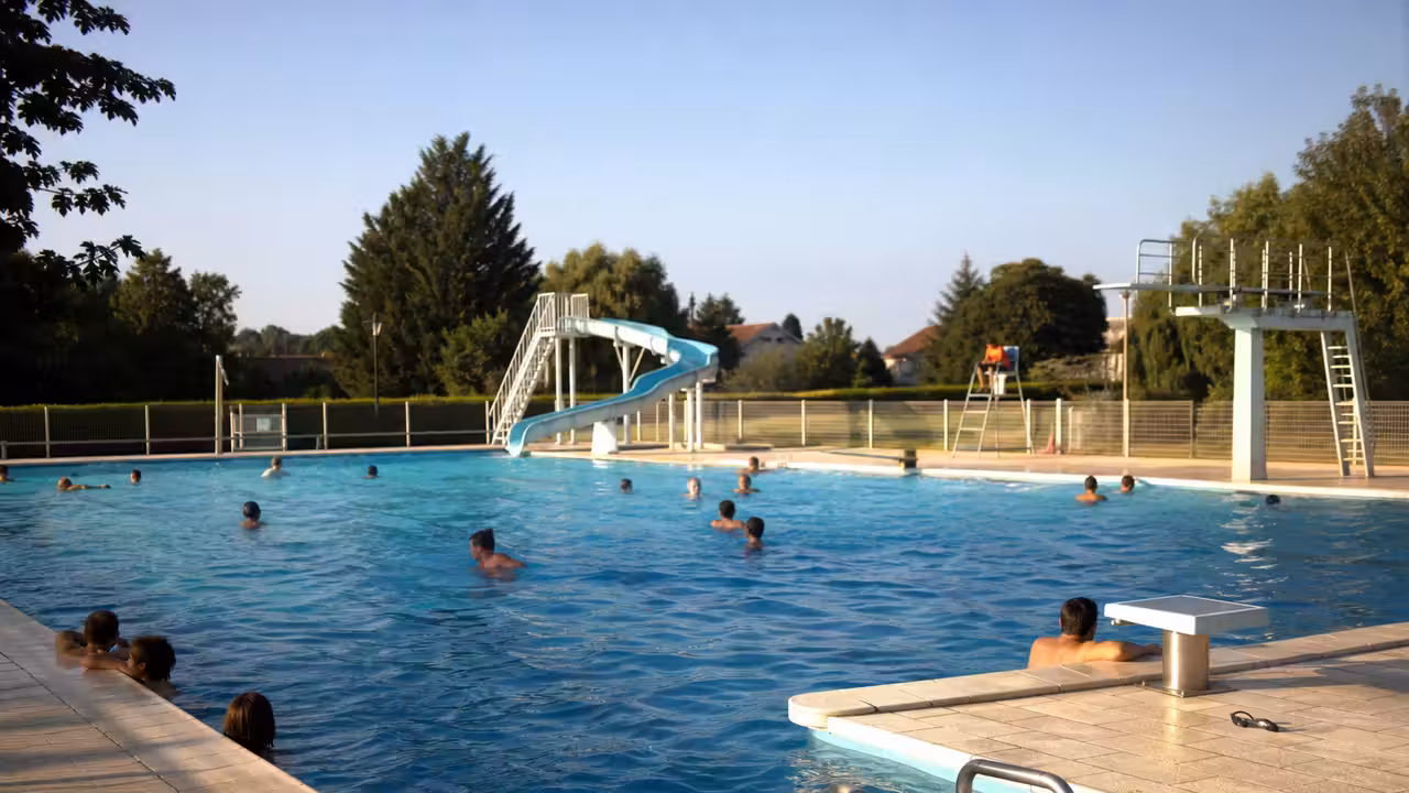 Bassin extérieur d’une piscine municipale avec toboggan, plages minérales et baigneurs vus de loin en été