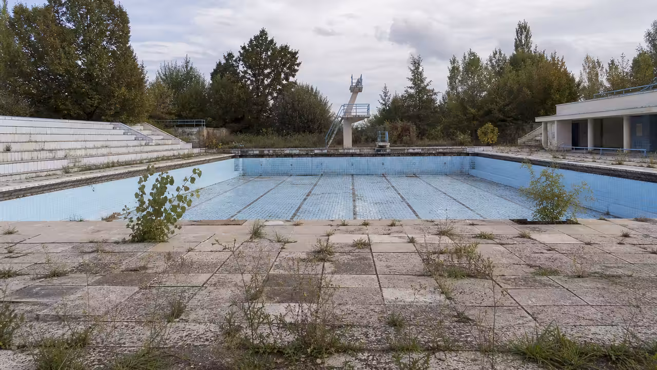 Ancien bassin extérieur vide, carrelage usé et végétation, évocation d'une piscine municipale fermée