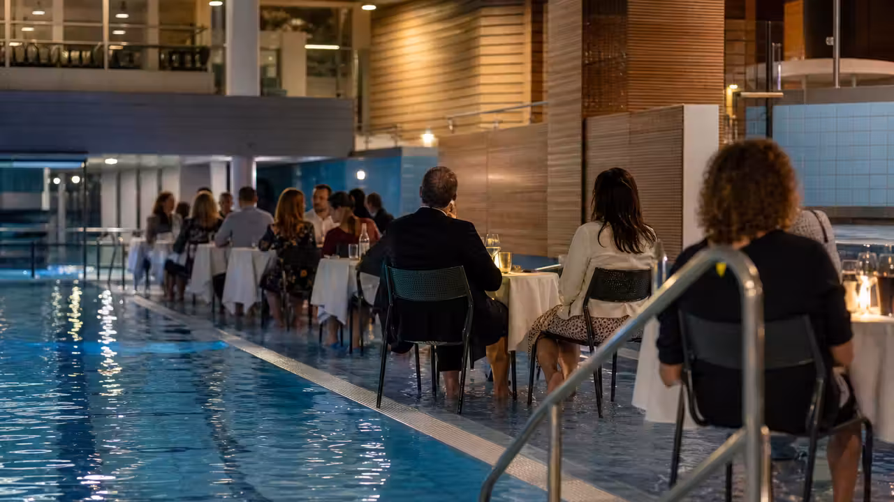 Table dressée au bord d’un bassin intérieur, avec des convives assis les pieds dans une eau peu profonde