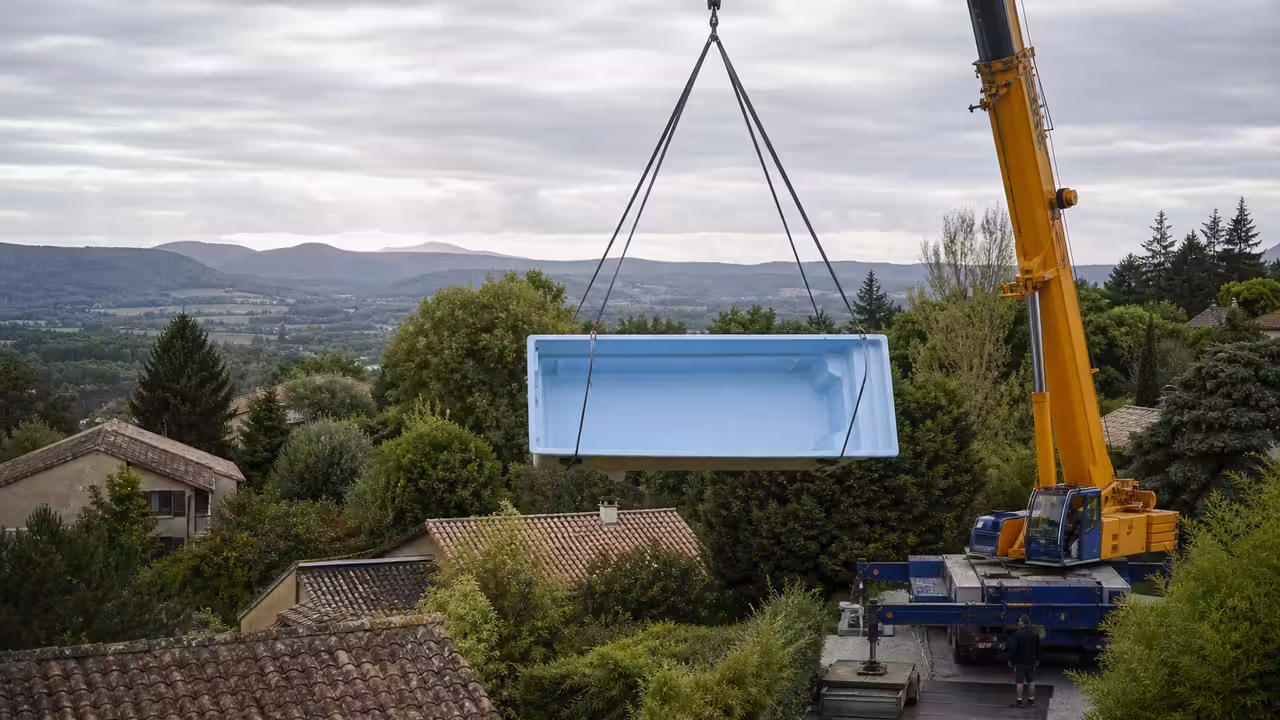 Une piscine coque suspendue par une grue au-dessus d'un jardin en pente entre des maisons.