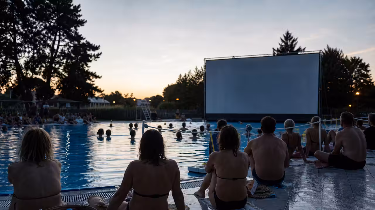 Projection nocturne sur grand écran au bord d’un bassin public éclairé, avec spectateurs en maillot de bain