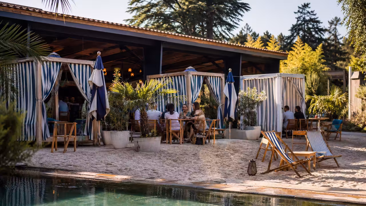 Terrasse de guinguette estivale avec bassin, transats, sable et cabanes rayées dans une ambiance balnéaire.